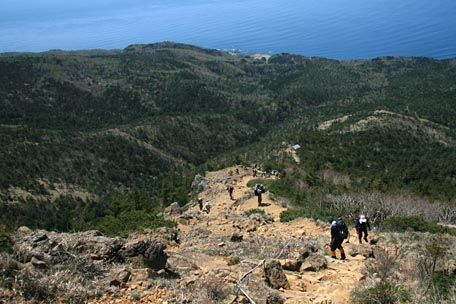 かんらん岩の赤茶けた登山道を登ってくる登山者とその向こうに海を望んだ写真